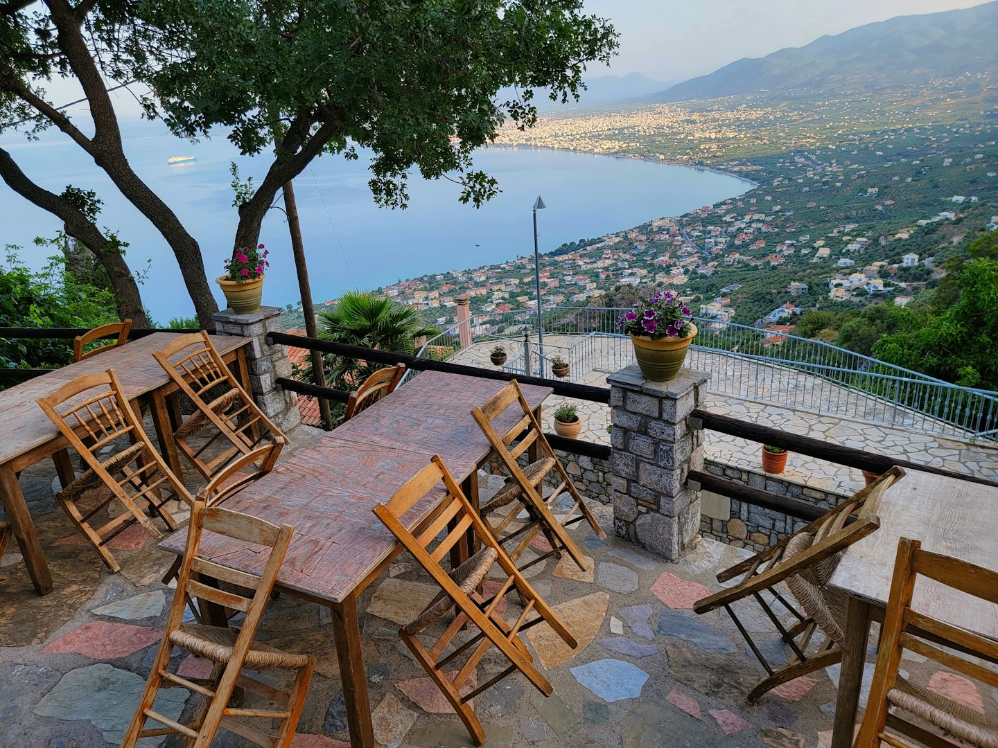 Wooden tables and chairs on a stone patio overlooking a coastal town.